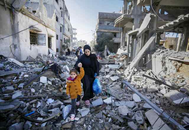 Ein kleines Kind in einer gelben Jacke hält die Hand eines Erwachsenen, während sie durch ein zerstörtes Viertel in Gaza gehen.
